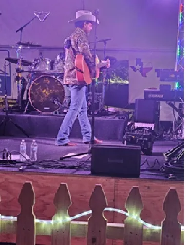 A country musician in a cowboy hat playing guitar on stage under stage lights, with a Texas flag hanging behind the drum kit.