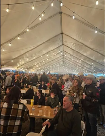 A packed event tent strung with lights, full of people in cowboy hats eating and talking around long tables.
